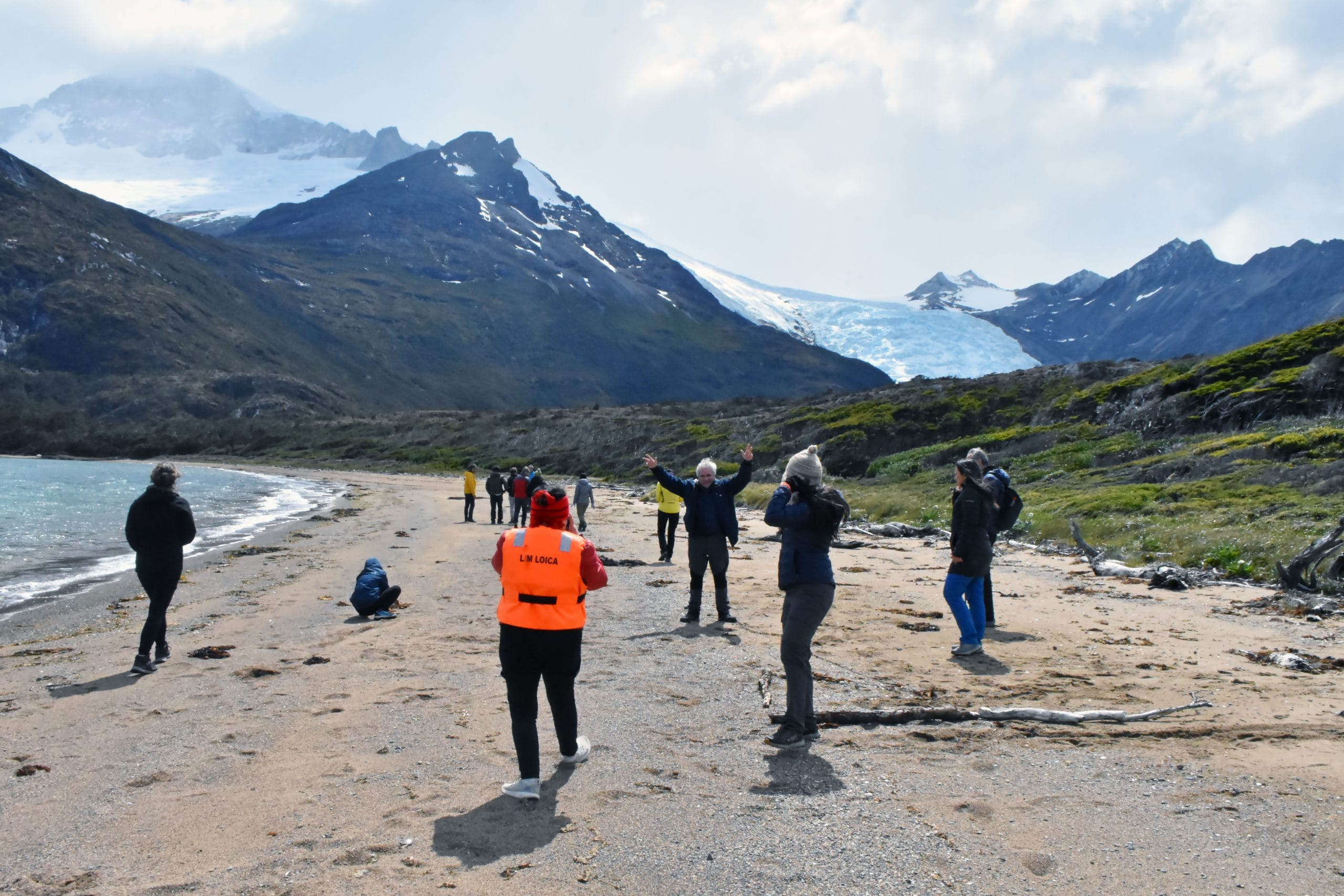 Grupo caminando por una playa al pie de las montañas, con un glaciar al fondo.