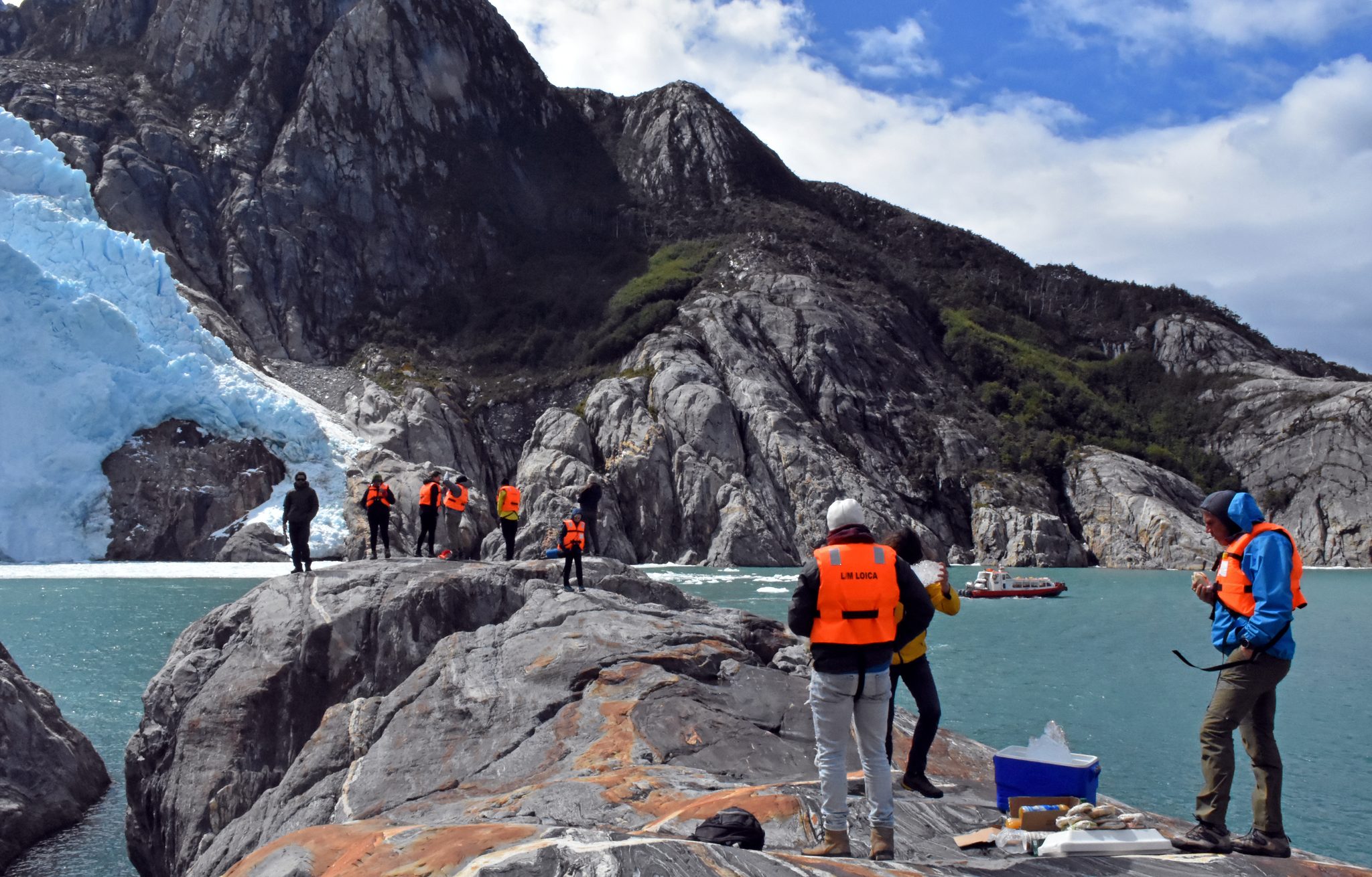 Visitantes con chalecos salvavidas sobre una roca al pie del glaciar, con la embarcación LOICA al fondo.