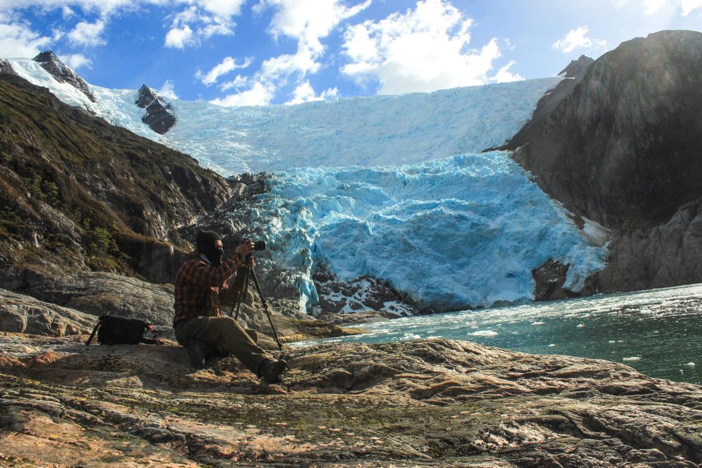 Un fotógrafo con trípode frente al frente del glaciar, con hielo azul y témpanos en el agua.