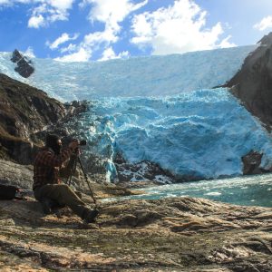 Un fotógrafo con trípode frente al frente del glaciar, con hielo azul y témpanos en el agua.