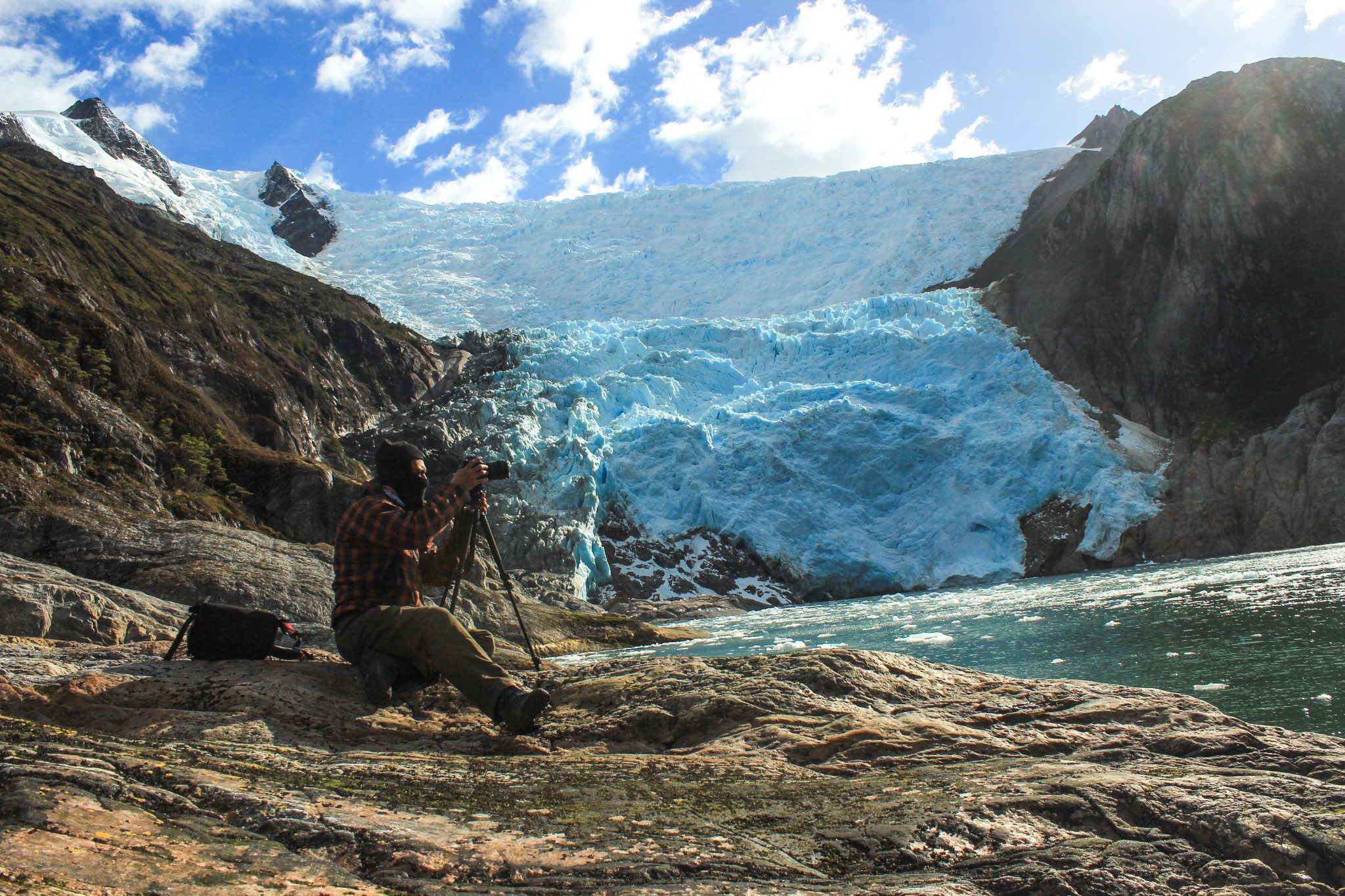 Un fotógrafo con trípode frente al glaciar, con hielo azul y témpanos en el agua.