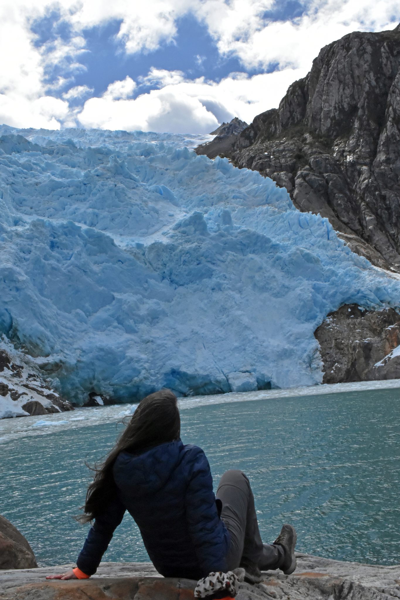 Una visitante sentada en la roca frente al frente de hielo del glaciar y el agua turquesa.