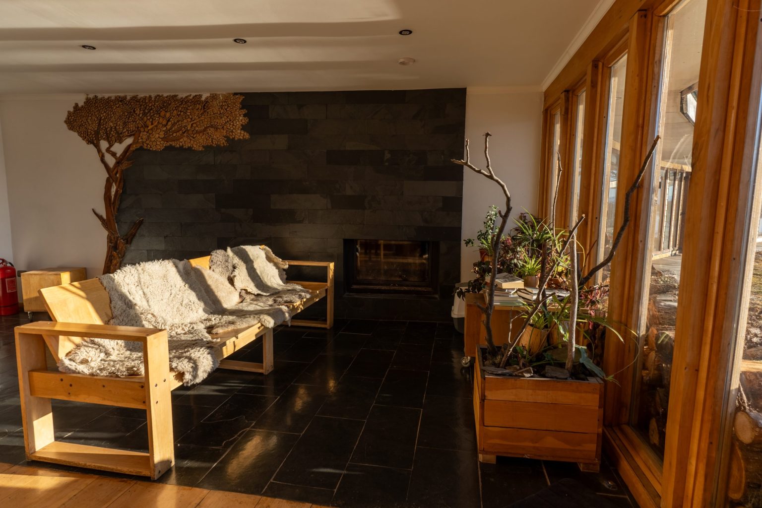 Living del lodge con chimenea, sillones de madera cubiertos con pieles y plantas junto al ventanal.