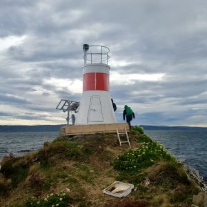 Un faro blanco y rojo sobre una punta cubierta de pasto, con el canal y un cielo cargado de nubes.
