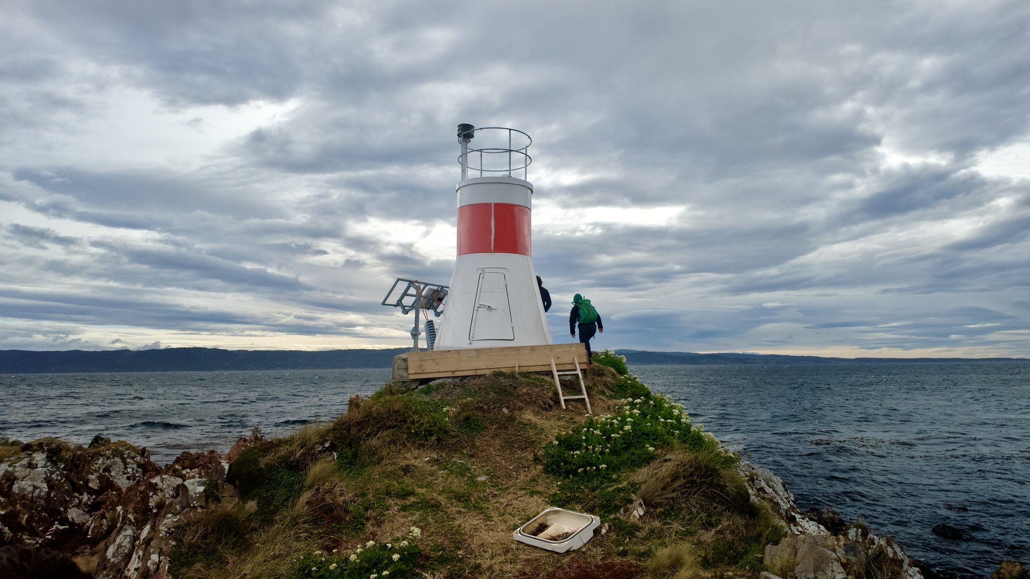 Un faro blanco y rojo sobre una punta cubierta de pasto, con el canal y un cielo cargado de nubes.